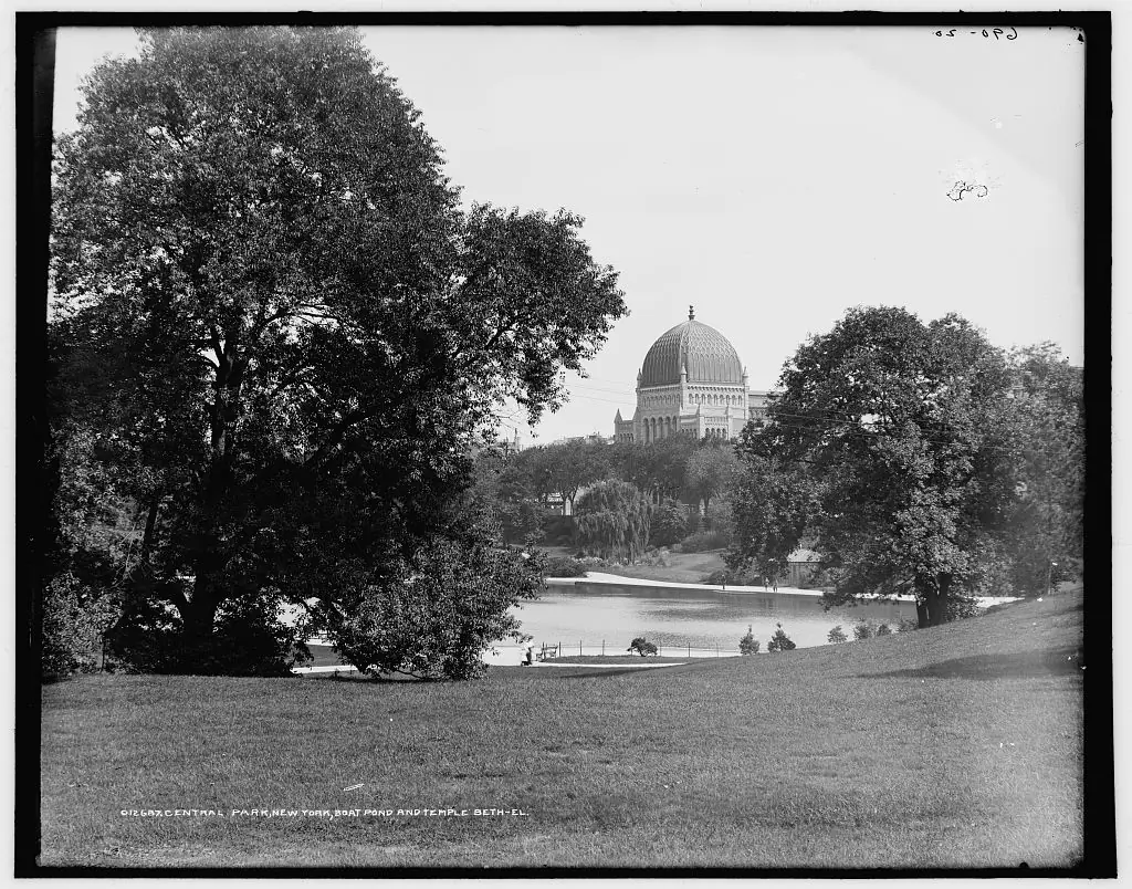 Vista histórica del estanque de Central Park entre árboles, con Temple Beth-El al fondo