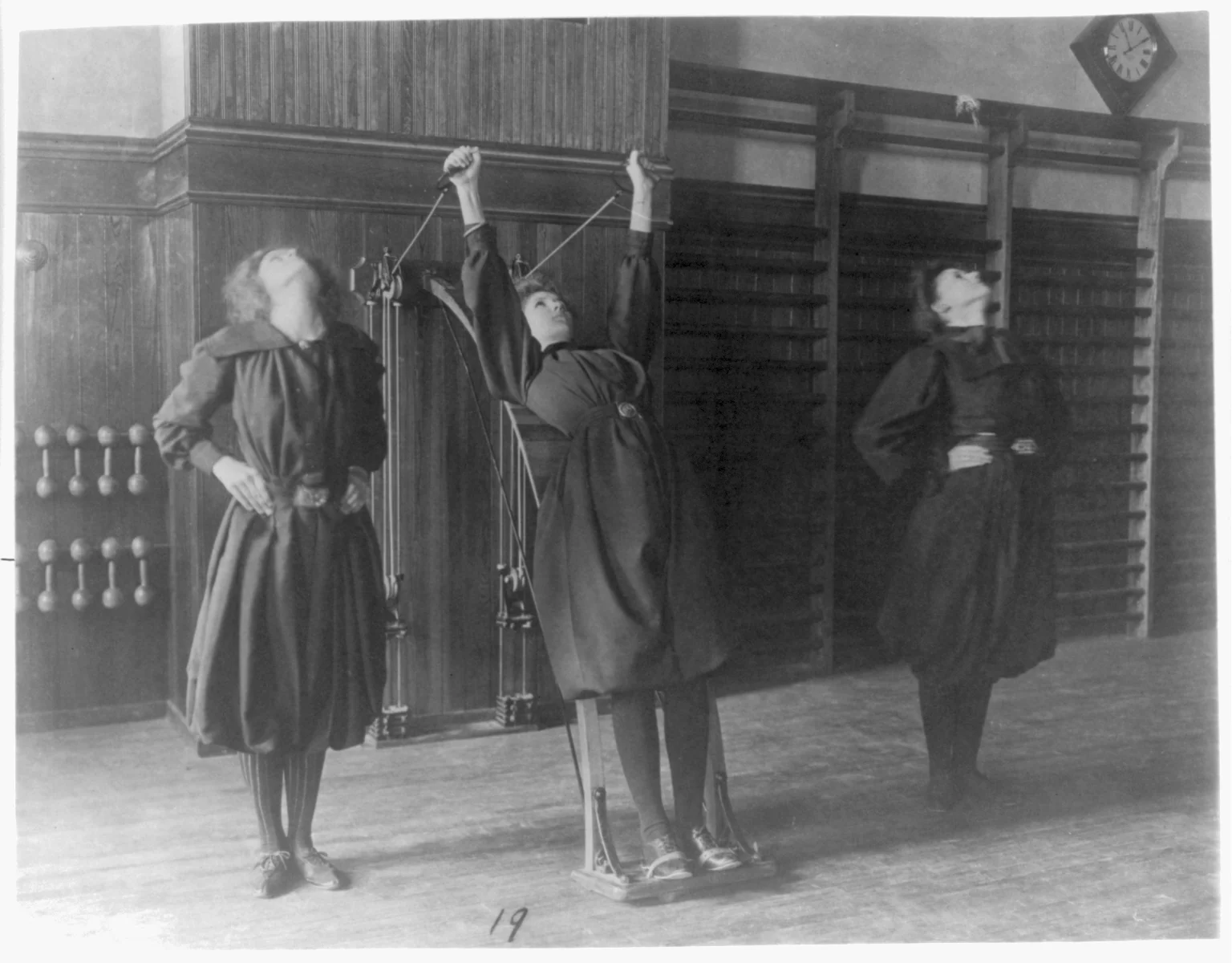 Tres estudiantes en un gimnasio histórico; una utiliza un aparato de cuerdas y poleas fijado a la pared.
